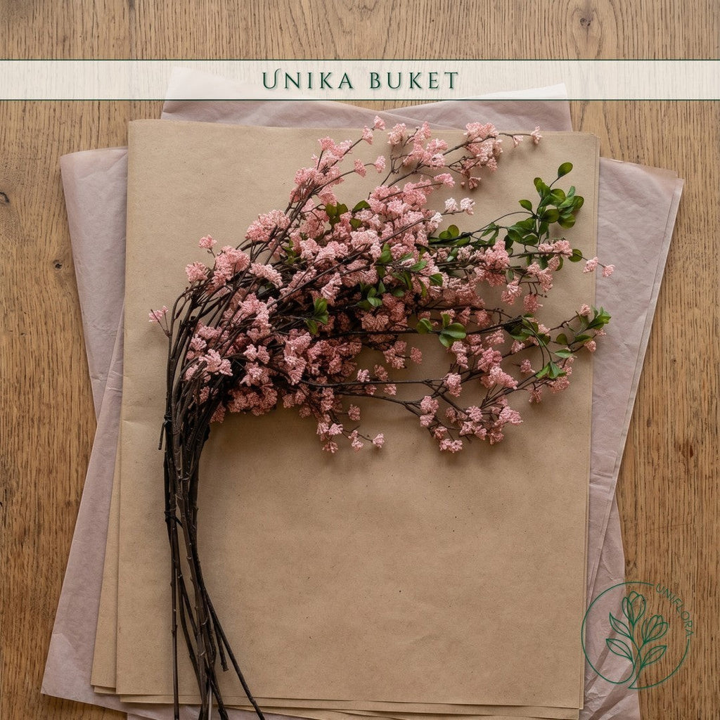 Uniflora Unika 26.60 buket med lyserøde kirsebærblomster på mørke grene og grønne blade, pakket i brunt papir.